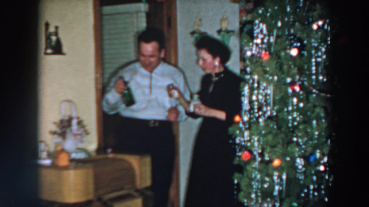Couple stands near a Christmas tree while the man holds a bottle and glass.