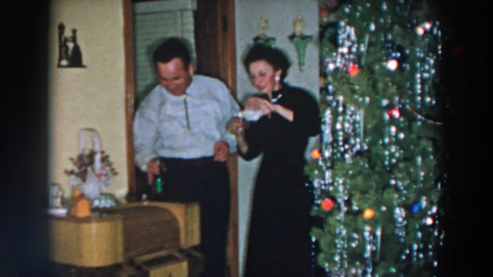 A woman drinks from a glass as a man stands nearby with a bottle, both beside a tinsel-trimmed Christmas tree in the home.