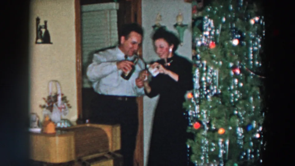 The man appears to pour or handle a drink while the woman smiles beside him near the decorated Christmas tree.