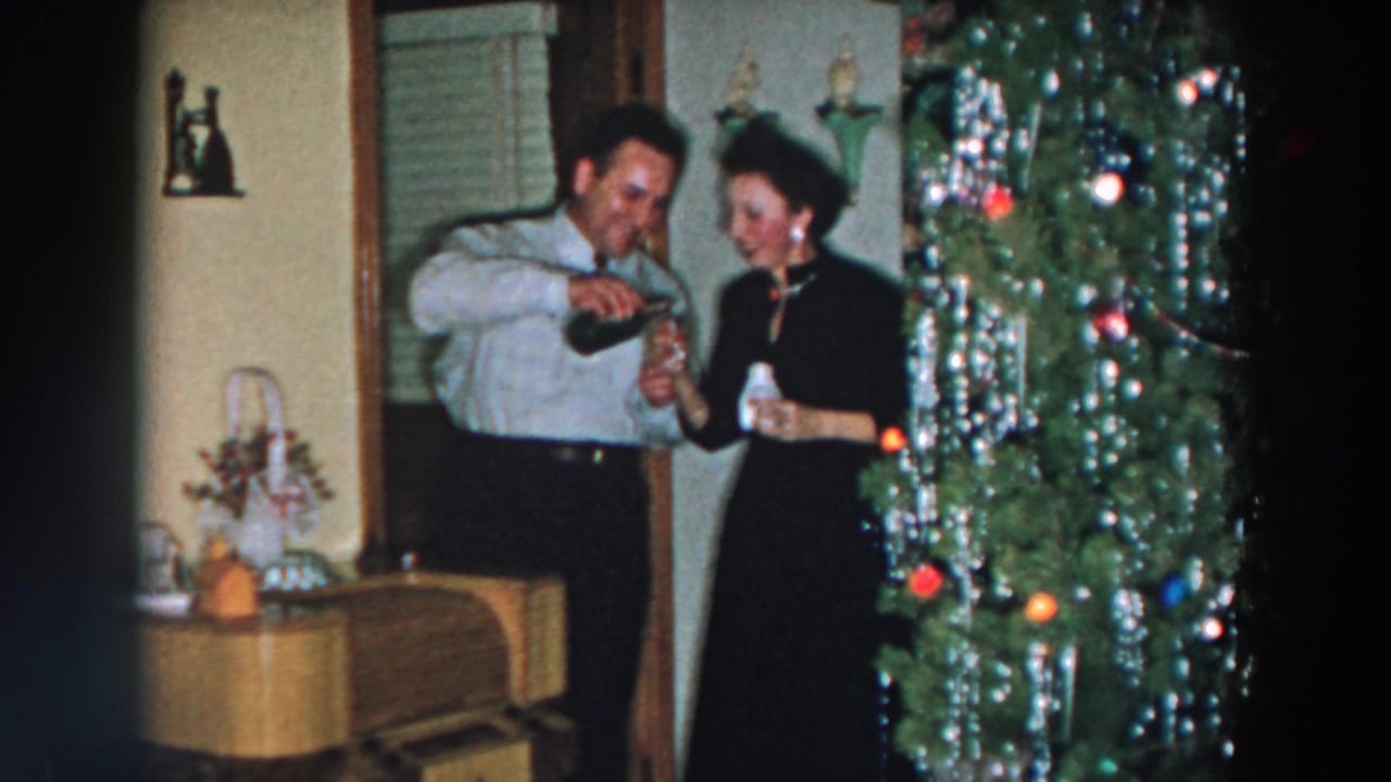 Man pours a drink for a woman beside a decorated Christmas tree in a home interior.