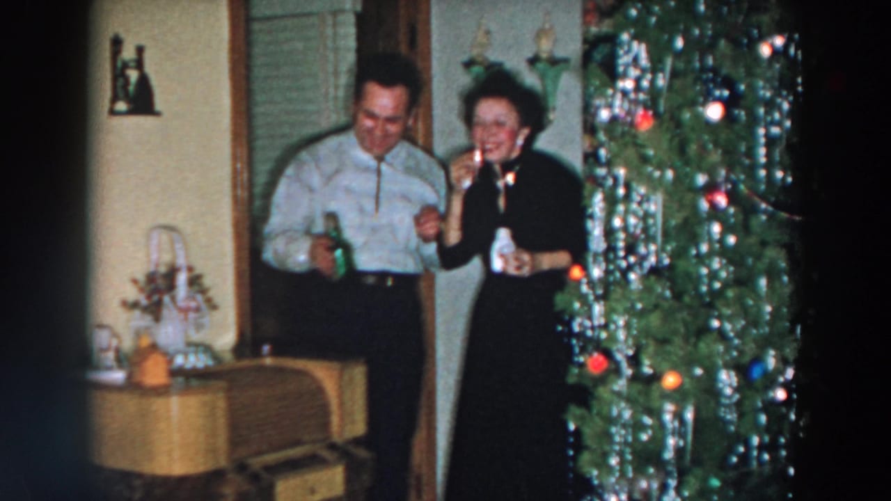 Man and woman hold drinks and smile near a Christmas tree with tinsel and ornaments.