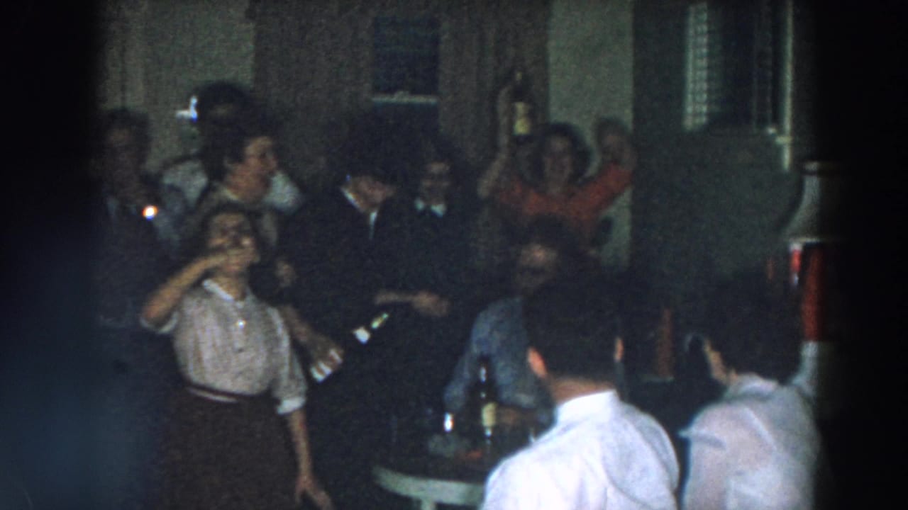 Adults gather in a dim room while a woman raises a bottle near seated guests.