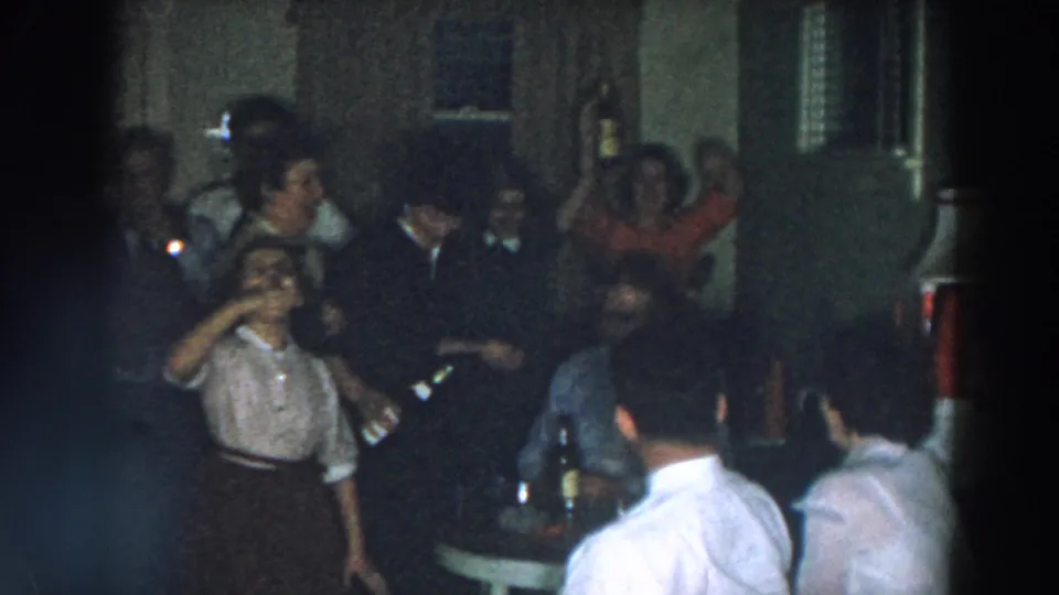 1957 home-movie frame from Wheeling, Ohio showing adults in a dim private interior with a woman raising a bottle during a party.