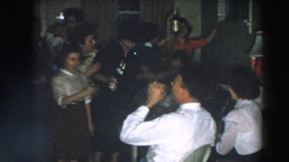 A handheld 1957 Wheeling, Ohio party frame shows adults seated and standing close together, with bottles lifted in celebration.