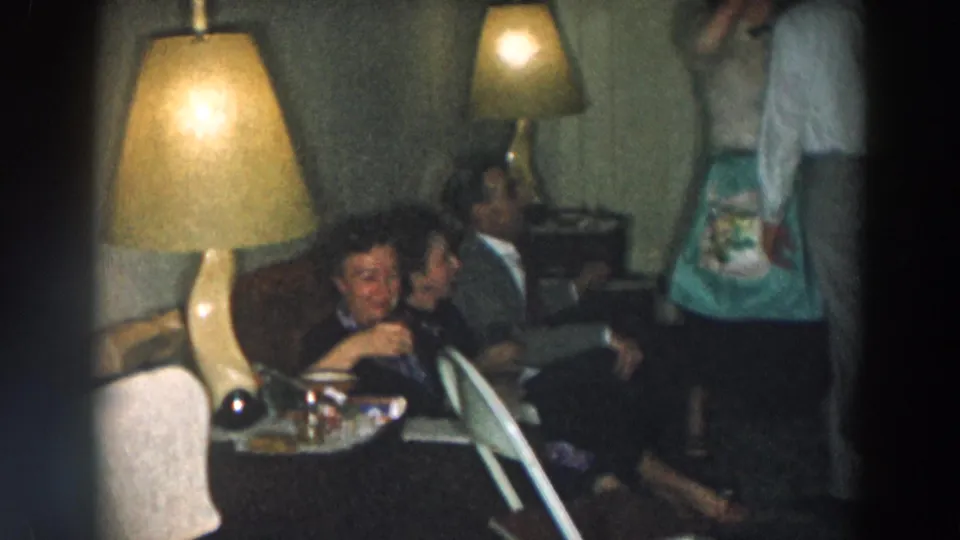 1957 Wheeling, Ohio home-movie frame of adults gathered on a couch in a lamp-lit private living room.
