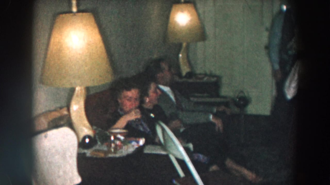 Woman drinks from a glass while seated with another adult on a couch.