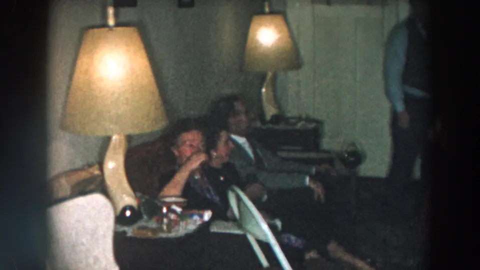 Adults share a couch in a softly focused 1957 Wheeling, Ohio home-movie frame with lamps and party tableware nearby.