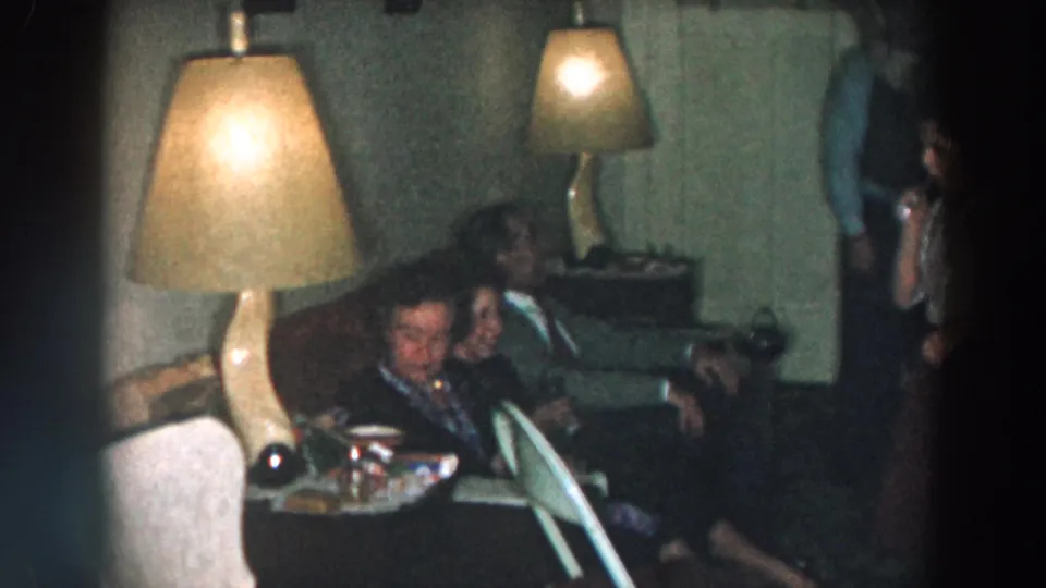 The home-movie frame shows seated adults and a partially visible standing person in a 1957 Wheeling, Ohio private interior.