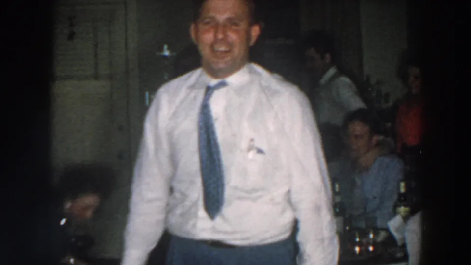 A smiling man in a white shirt and blue tie faces the camera inside the 1957 Wheeling, Ohio party scene, with guests behind him.
