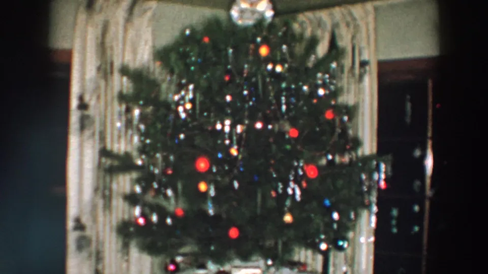 1957 Wheeling, Ohio frame of a lit Christmas tree with reflective tinsel, red ornaments, and a visible tree topper inside a private home.