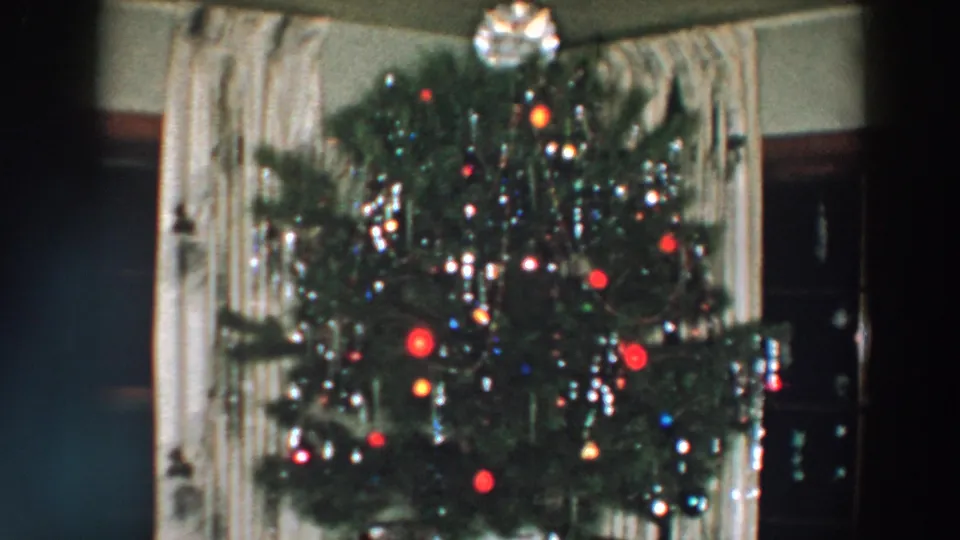 1957 Wheeling, Ohio home-movie frame of a decorated Christmas tree with hanging tinsel, colored lights, red ornaments, and a private interior background.
