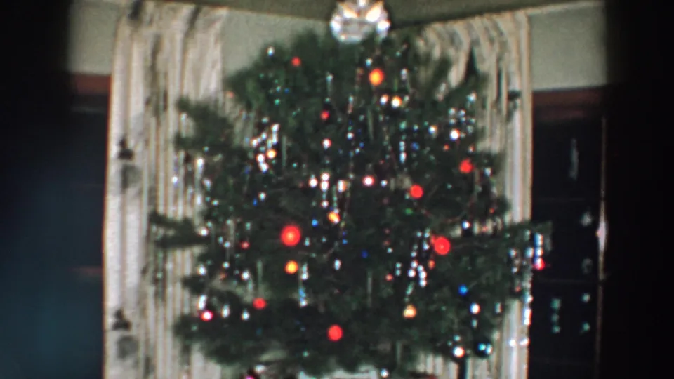 1957 Wheeling, Ohio home-movie frame showing a decorated Christmas tree with colored bulbs, tinsel, ornaments, curtains, and dark furniture.