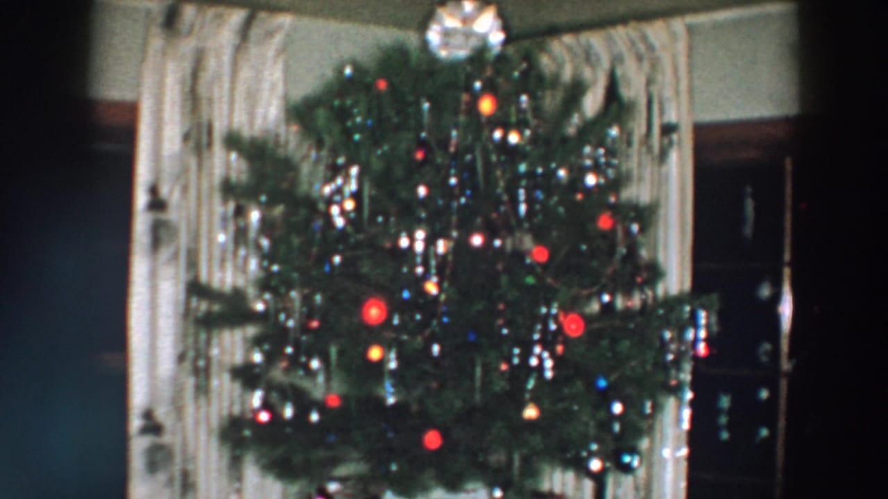 1957 Wheeling, Ohio frame of a Christmas tree with bright colored bulbs, silver tinsel, ornaments, curtains, and dark furniture nearby.