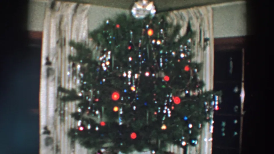 1957 Wheeling, Ohio frame of a Christmas tree with bright colored bulbs, silver tinsel, ornaments, curtains, and dark furniture nearby.