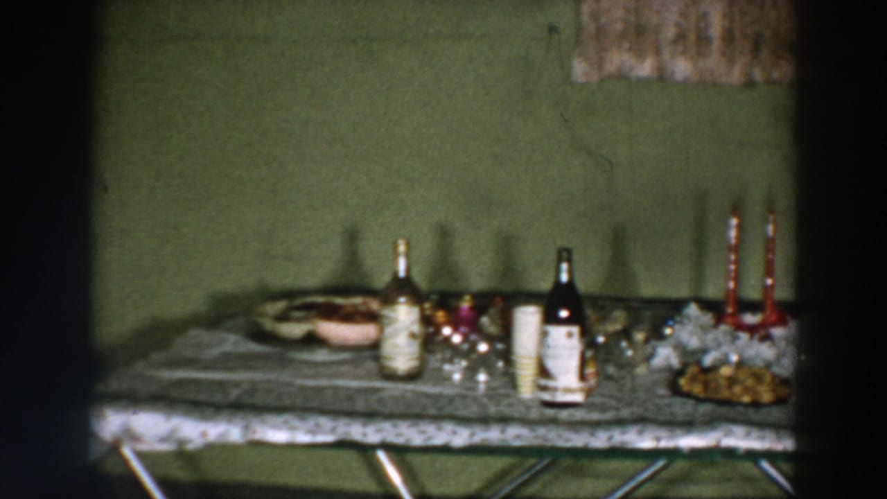 1957 Wheeling, Ohio home-movie frame showing a green wall and a party table with bottles, cups, snacks, glassware, and red taper candles.