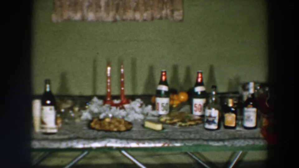 1957 Wheeling, Ohio home-movie frame showing a mid-century refreshment table with bottles, candles, white decoration, snacks, oranges, and cups.