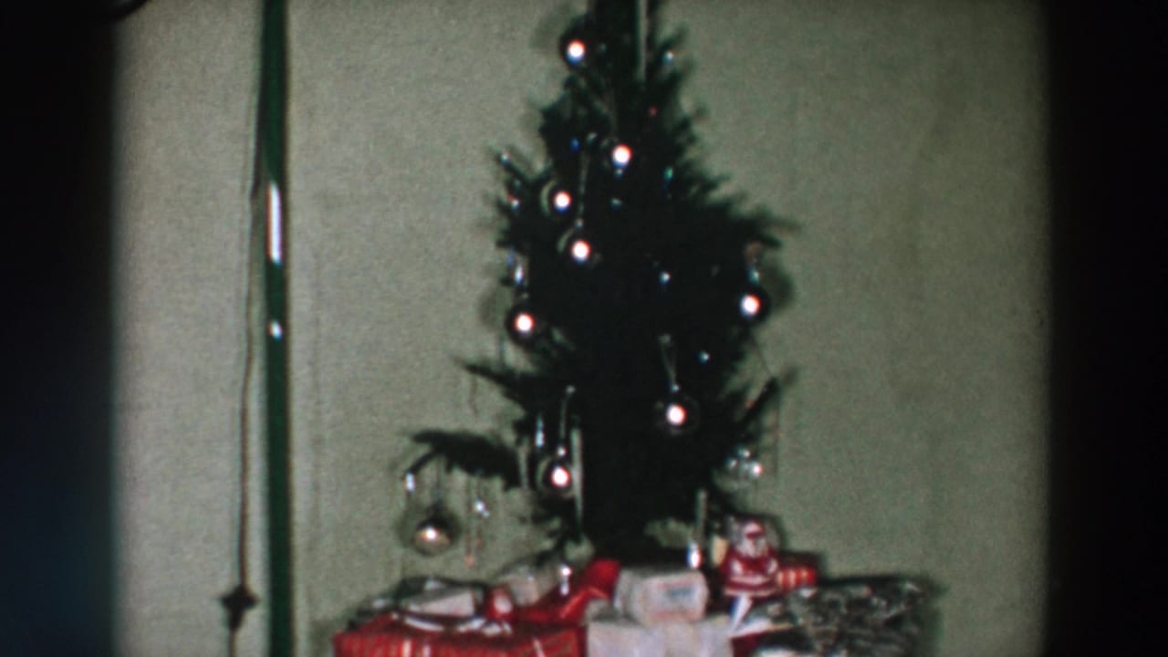 Small Christmas tree with ornaments beside wrapped gifts in a dim room.