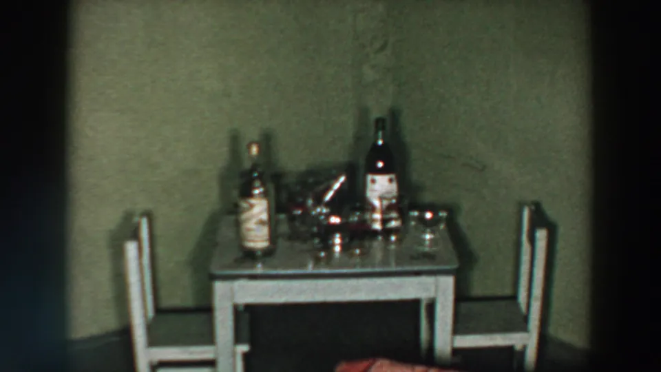 1957 Wheeling, Ohio home-movie frame showing a small table set with liquor bottles, glasses, and two chairs.