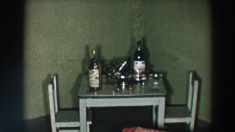 1957 Wheeling, Ohio home-movie frame showing a small table for two with liquor bottles, glasses, and a shaker against a plain wall.
