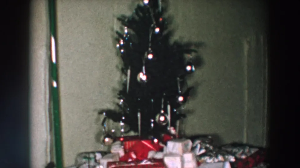 1957 Wheeling, Ohio frame of a Christmas tree with shiny ornaments, lights, and wrapped presents in a private room.