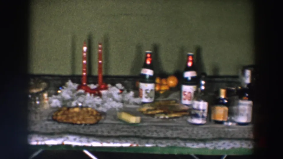 1957 Wheeling home-movie frame showing a refreshment table with red candles, bottles, snacks, oranges, jars, and a patterned cloth.