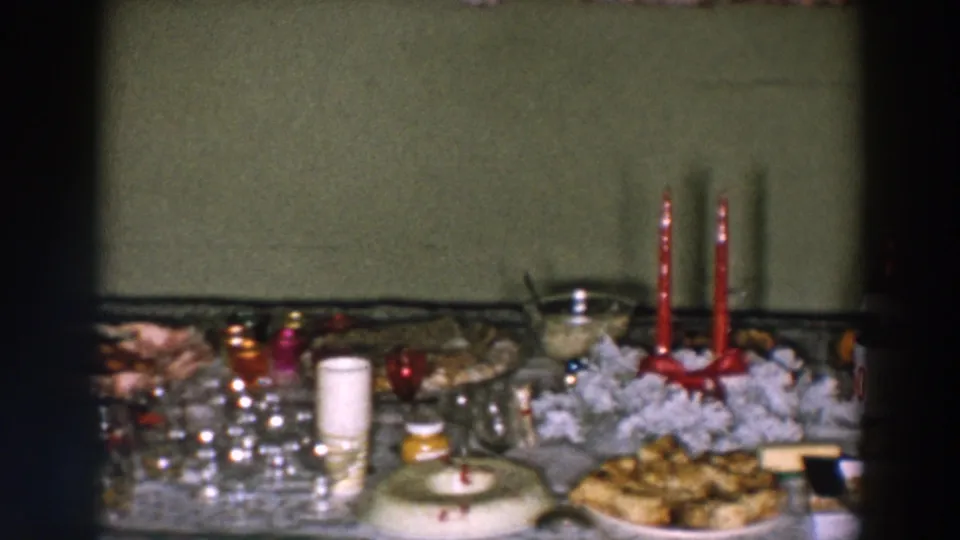 1957 Wheeling home-movie frame of a party table with red candles, plates of food, glassware, serving bowls, and assorted snacks.