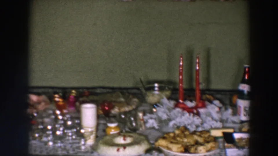 1957 Wheeling, Ohio home-movie frame showing a festive table with food, glassware, red candles, bottles, and a white wreath-like centerpiece.