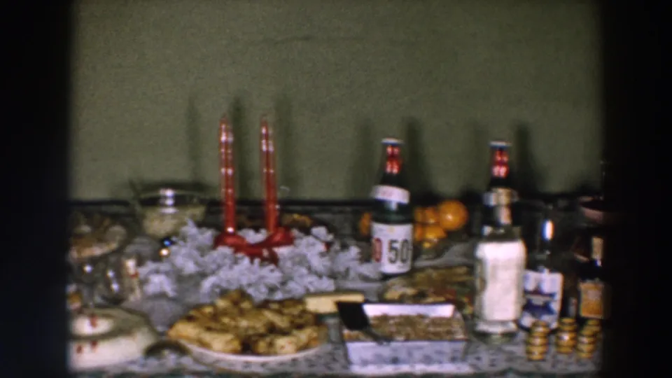 1957 home-movie frame showing desserts and white decoration around red candles, with labeled bottles and serving items nearby.