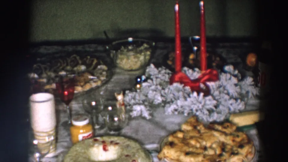 1957 Wheeling, Ohio home-movie frame showing a holiday dining spread with candles, wreath decor, glassware, a cake, pastries, and a small labeled bottle.