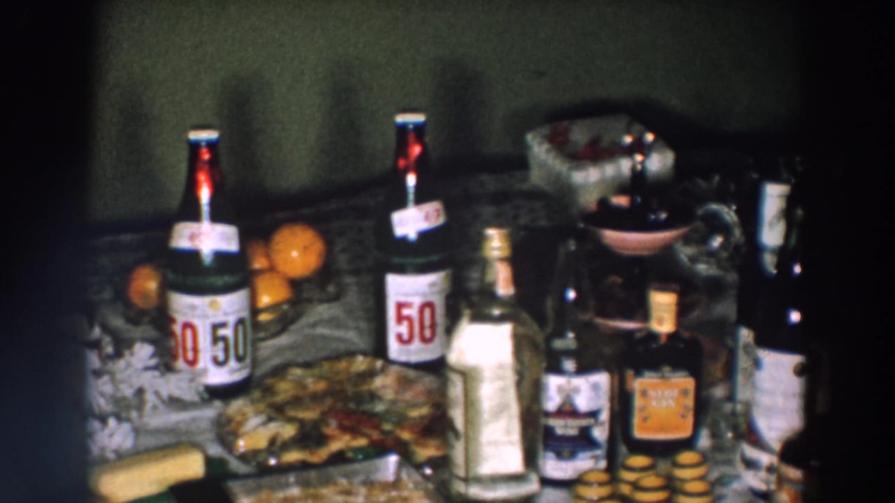 1957 Wheeling, Ohio home-movie frame showing 50/50 soda bottles, oranges, baked food, a clear bottle, and dark beverage bottles on a table.