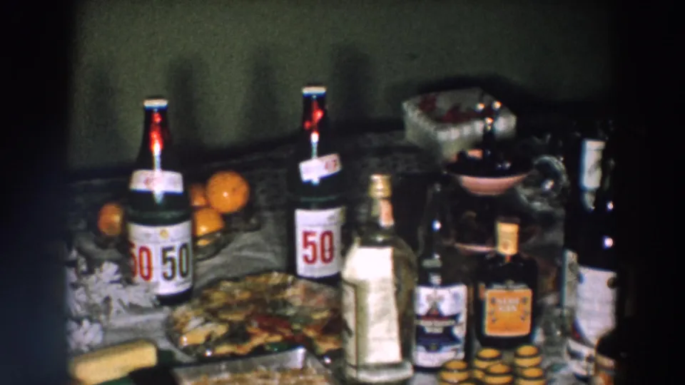 1957 Wheeling, Ohio home-movie frame showing 50/50 soda bottles, oranges, baked food, a clear bottle, and dark beverage bottles on a table.