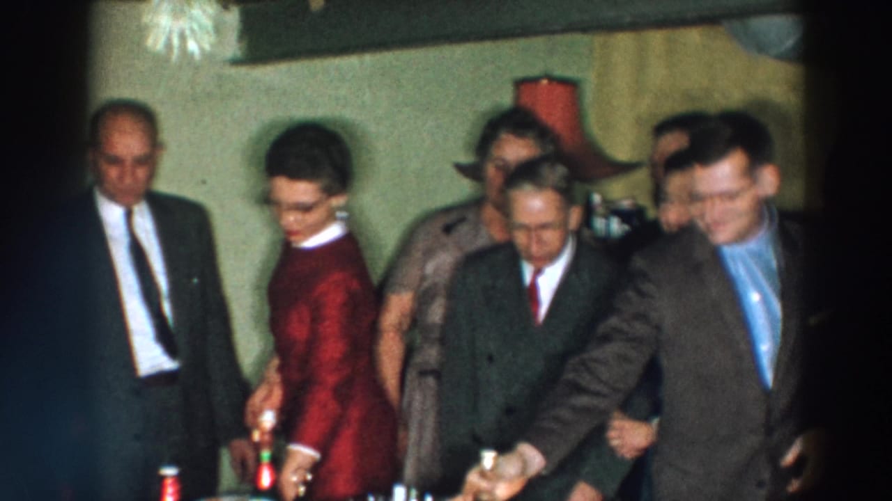 1957 Wheeling, Ohio home-movie frame showing formally dressed adults near a refreshment table with bottles and small glasses.