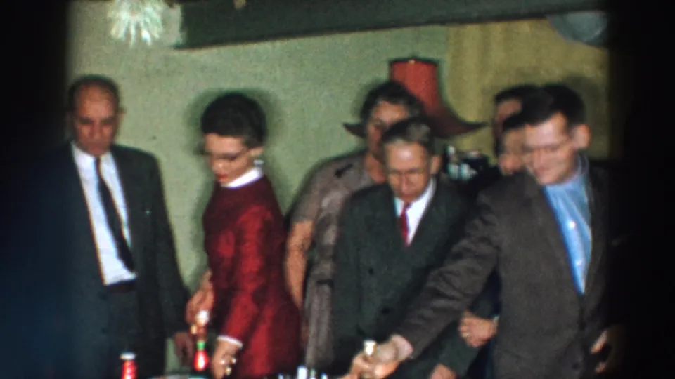 1957 Wheeling, Ohio home-movie frame showing formally dressed adults near a refreshment table with bottles and small glasses.
