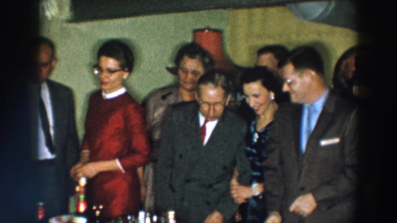 1957 Wheeling, Ohio frame with adults in suits and formal dresses standing closely by a table with bottles and glassware.