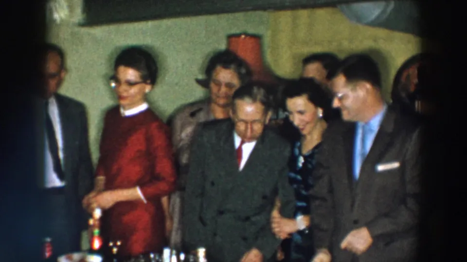 1957 Wheeling, Ohio frame with adults in suits and formal dresses standing closely by a table with bottles and glassware.