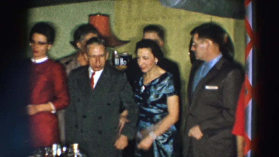 1957 Wheeling, Ohio frame of a formal indoor group beside bottles, with a red-and-white striped vertical edge visible at right.