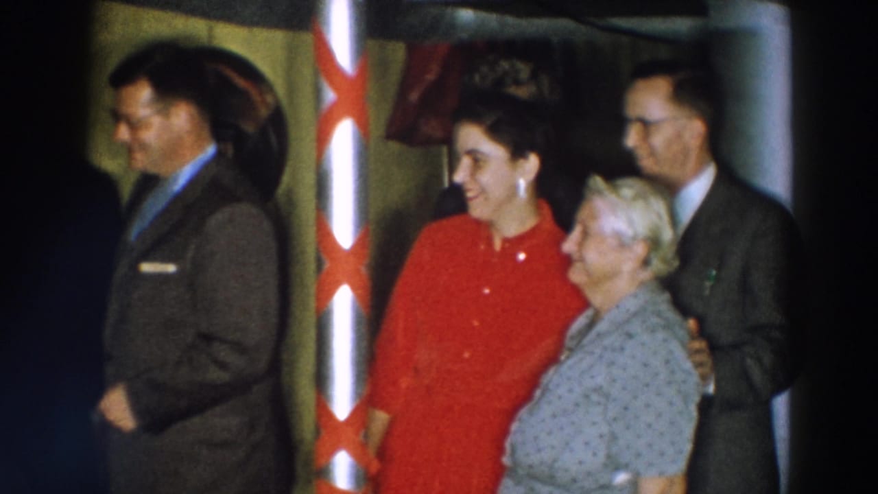 The 1957 Wheeling, Ohio frame shows a woman in red standing with an older woman near a ribboned pole, with suited men nearby.