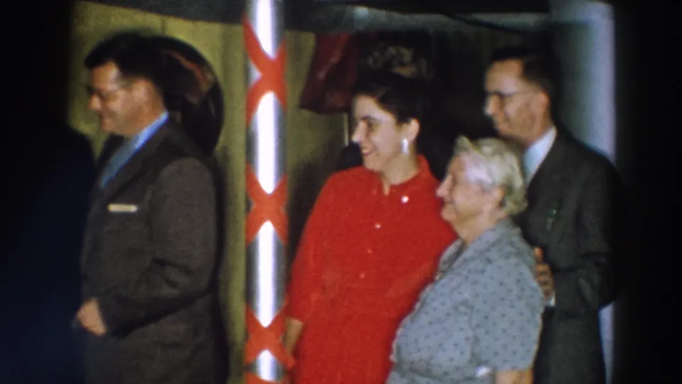 The 1957 Wheeling, Ohio frame shows a woman in red standing with an older woman near a ribboned pole, with suited men nearby.