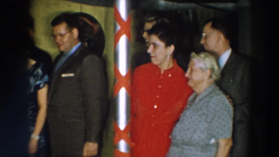 In this Wheeling, Ohio 1957 home-movie frame, adults in suits and dresses stand closely around a woman in red beside a ribboned pole.