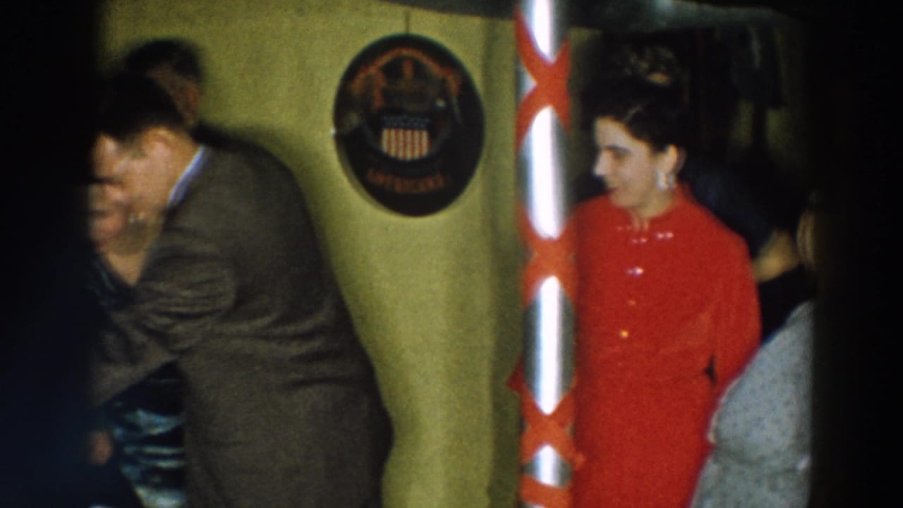 Adults in a warm interior, with a man leaning toward a woman and a woman in red standing nearby.