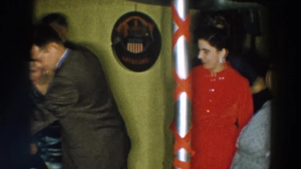 1957 Wheeling, Ohio home-movie frame showing adults gathered in a private room near a circular wall emblem and red lampshade.
