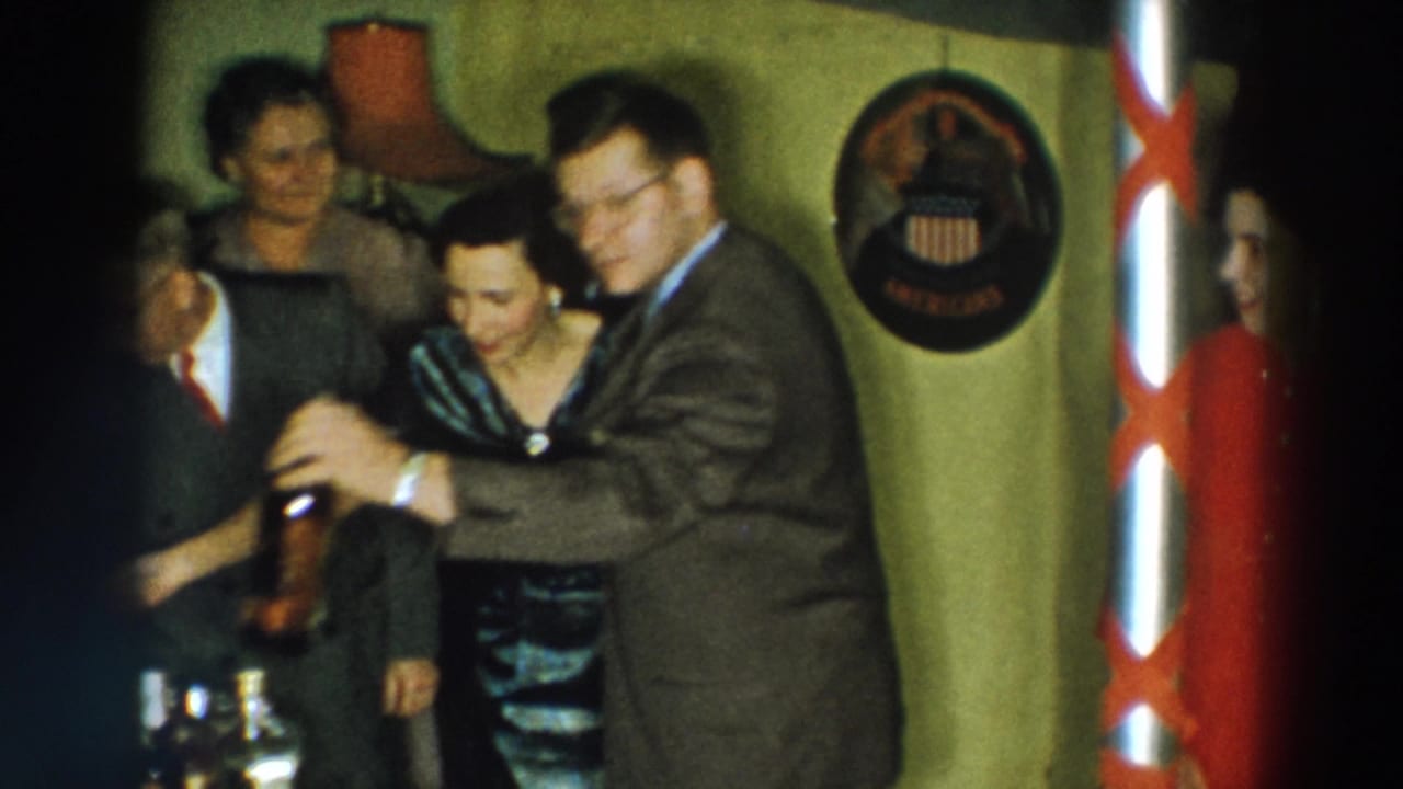 1957 Wheeling, Ohio home-movie frame of adults smiling and leaning together during a private indoor gathering.
