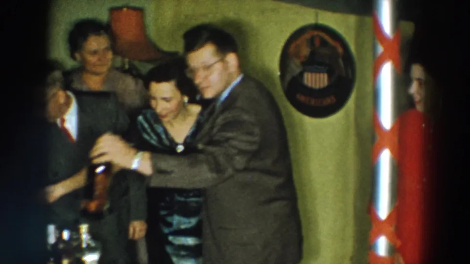 1957 Wheeling, Ohio home-movie frame of adults smiling and leaning together during a private indoor gathering.