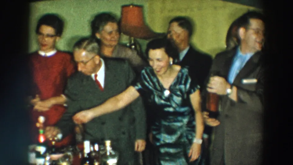 1957 Wheeling, Ohio home-movie frame showing adults clustered around drinks in a mid-century private room.