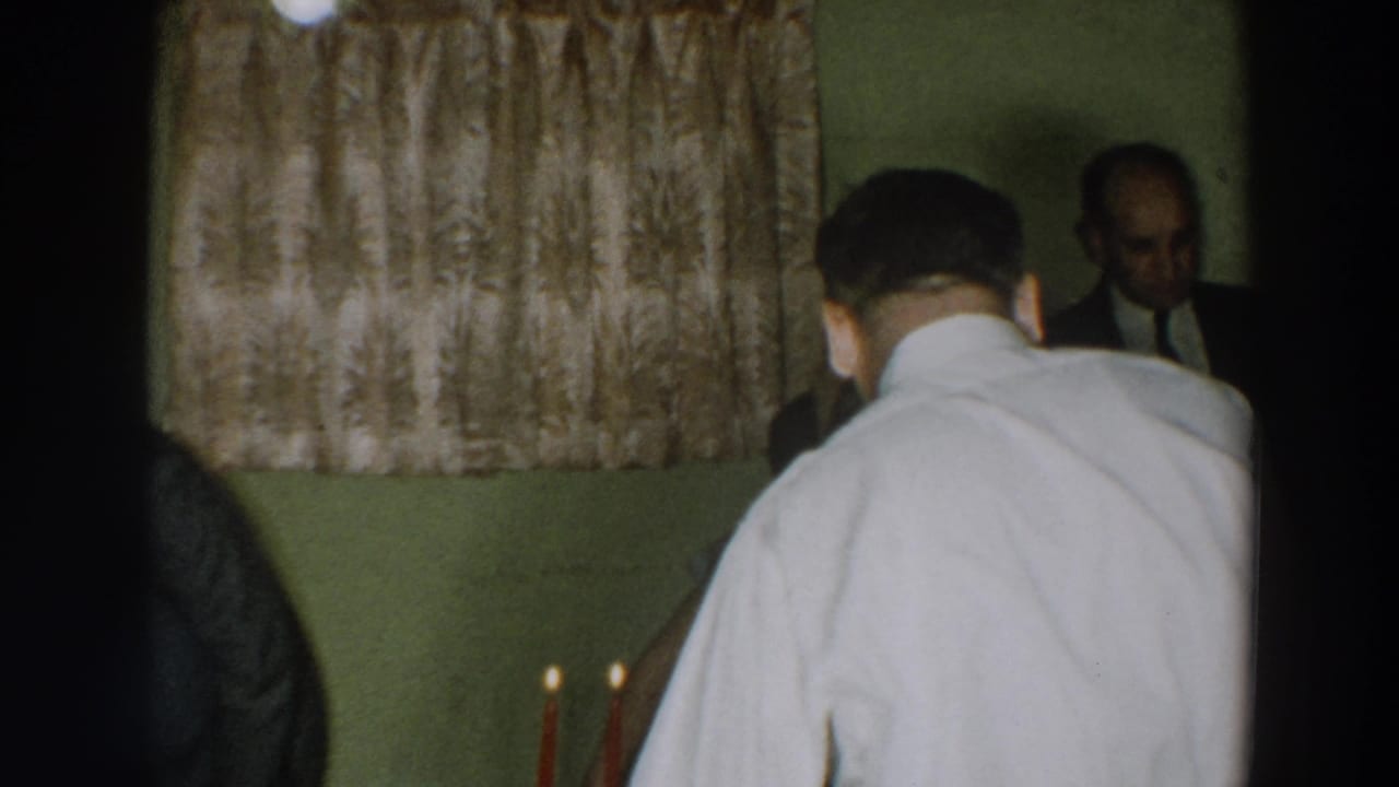 1957 home-movie frame from Wheeling, Ohio showing adults at an indoor party near patterned curtains and table candles.