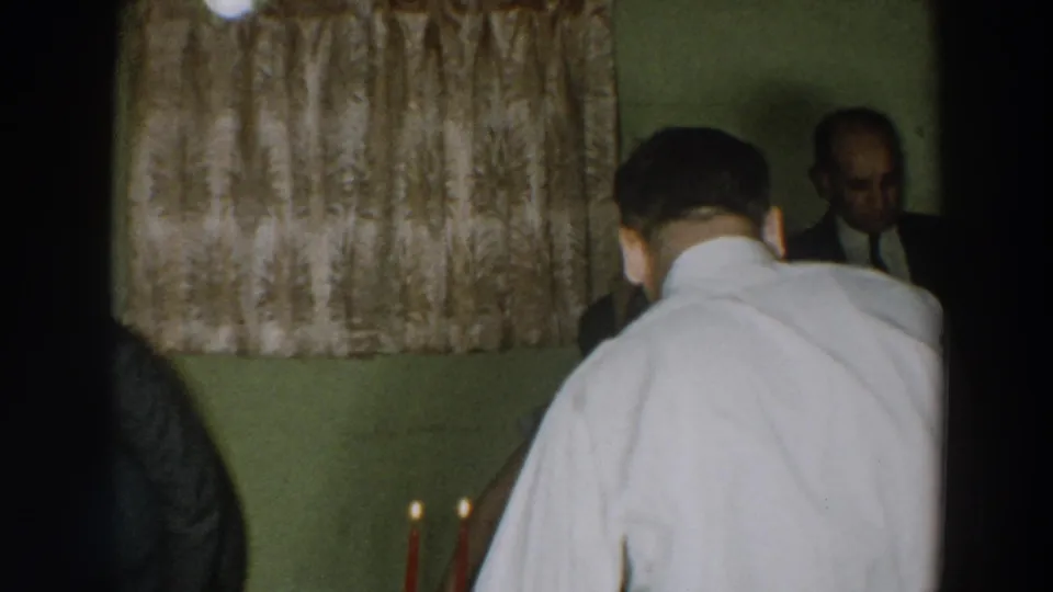 1957 home-movie frame from Wheeling, Ohio showing adults at an indoor party near patterned curtains and table candles.