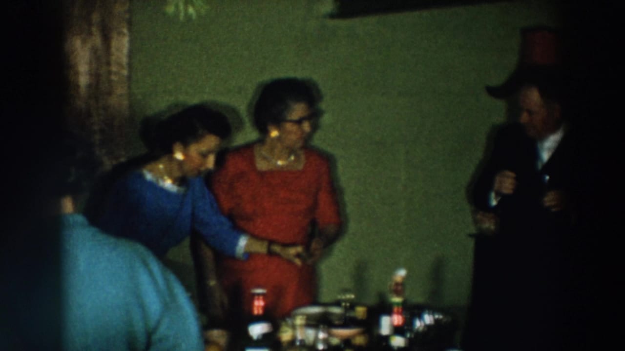 Women in blue and red dresses stand near a table while a man in a dark suit holds a drink.