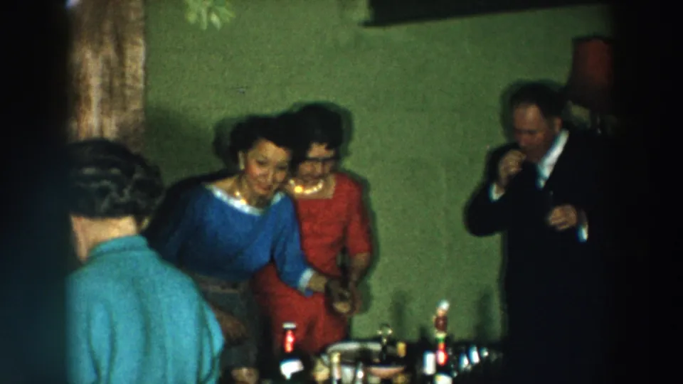 Home-movie frame of a 1957 Wheeling cocktail party with guests talking beside bottles, candles, and a lamp.
