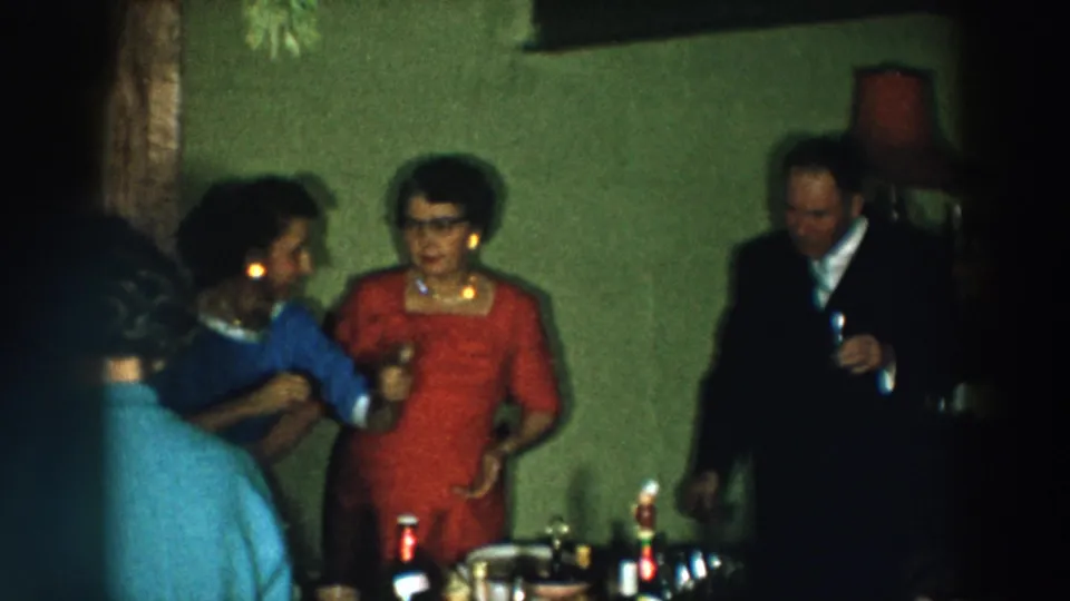 1957 Wheeling, Ohio indoor party frame with two women talking near a bottle-covered table and a suited man nearby.