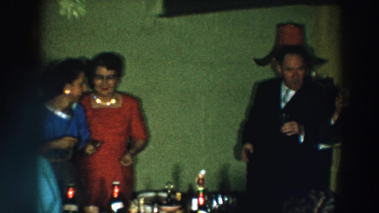 1957 Wheeling, Ohio home-movie frame showing adults in suits and dresses gathered near drinks in a private interior.
